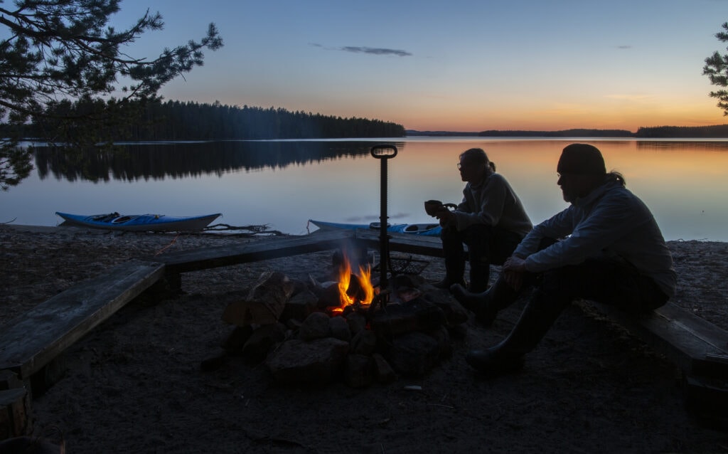 Nainen ja mies istuvat nuotion ääressä järven rannalla iltahämärässä